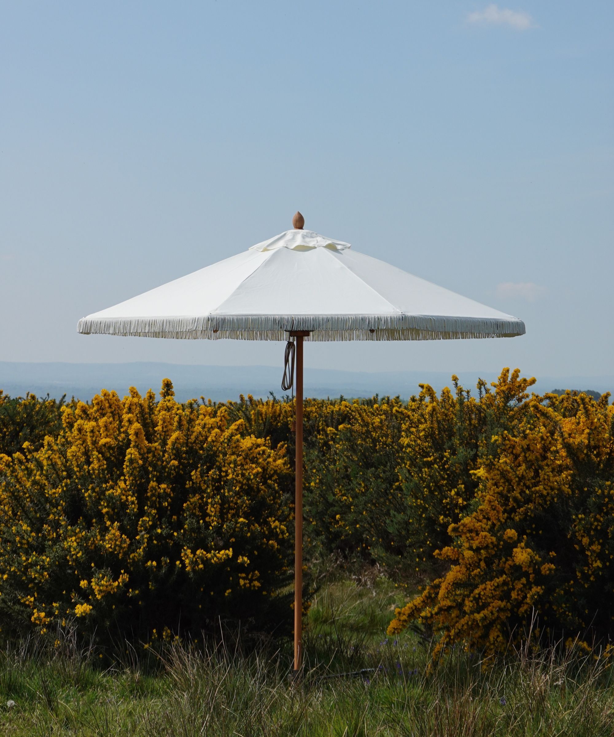 White parasol with tassels on a grass verge with a gorse bush behind