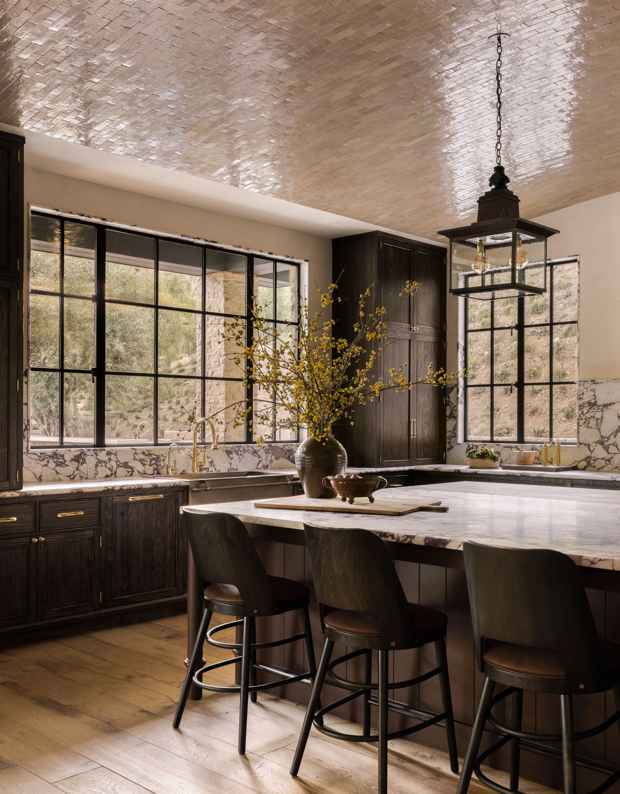 A modern kitchen with dark cabinetry and light worktops with a beige-colored tiled ceiling on the central arch, dark bar stools next to the large island, and a vase with yellow-flowered foliage on top