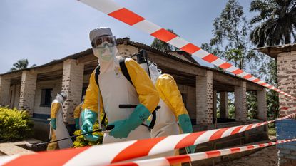 A series of people wearing yellow and white hazmat suits stand in front of a house behind an orange and white striped tape.