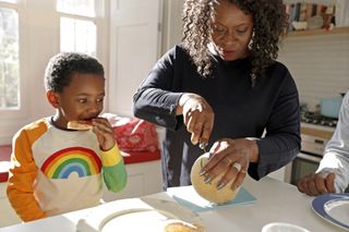 mother and son at home in the kitchen. Mother is cutting up a melon while son is eating lunch