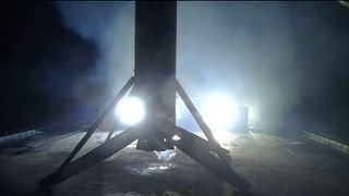 the first stage of a rocket is seen lit at night in silhouette standing on its four landing legs atop a sea-based droneship