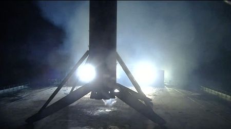 the first stage of a rocket is seen lit at night in silhouette standing on its four landing legs atop a sea-based droneship