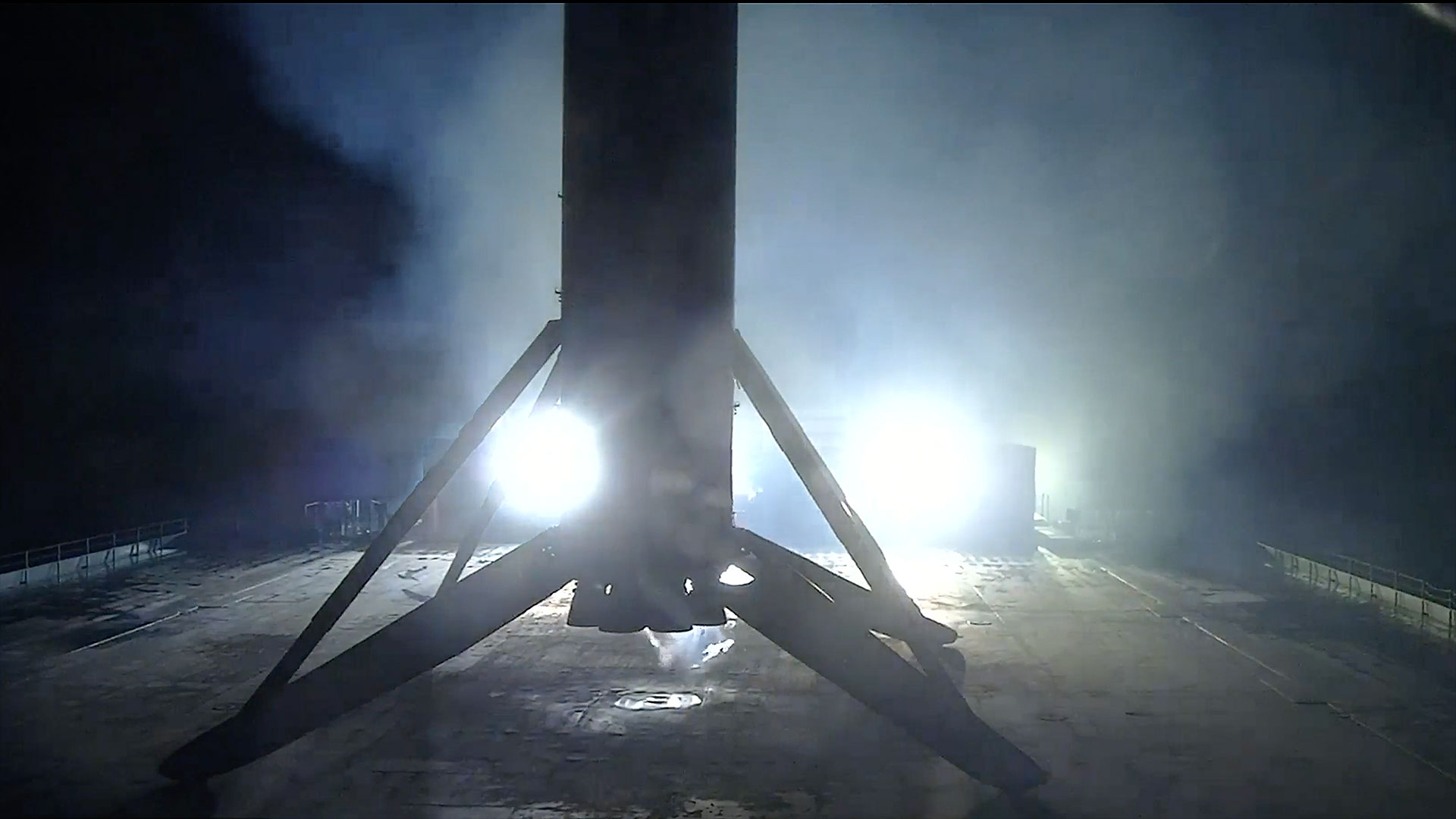 the first stage of a rocket is seen lit at night in silhouette standing on its four landing legs atop a sea-based droneship 