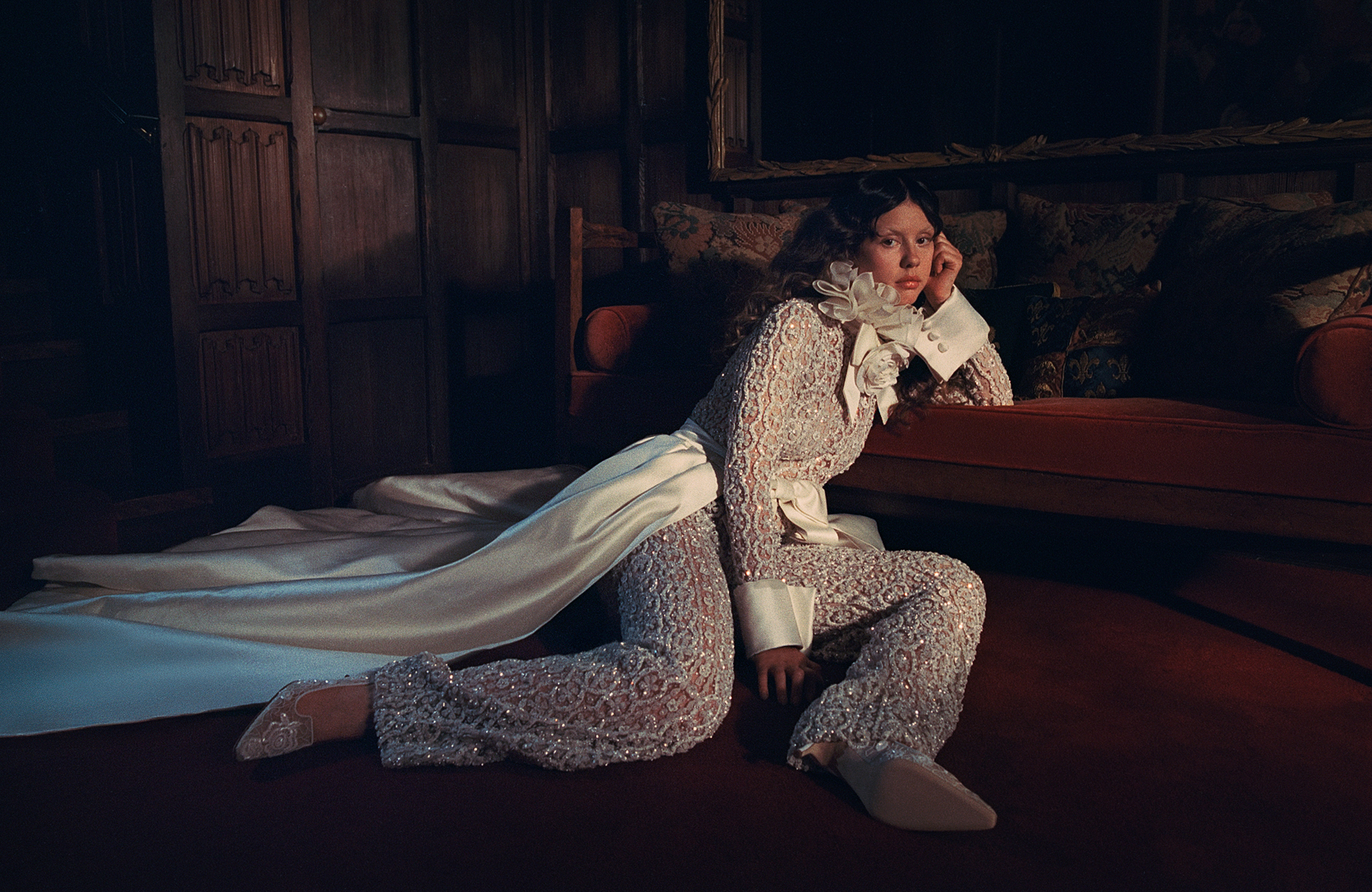 Mia Goth photo shoot for Who What Wear&#039;s October Cover Story. She is sitting on the floor, leaning on a velvet couch wearing a white crystal-embellished jumpsuit with satin bow detailing and ruffled collar.