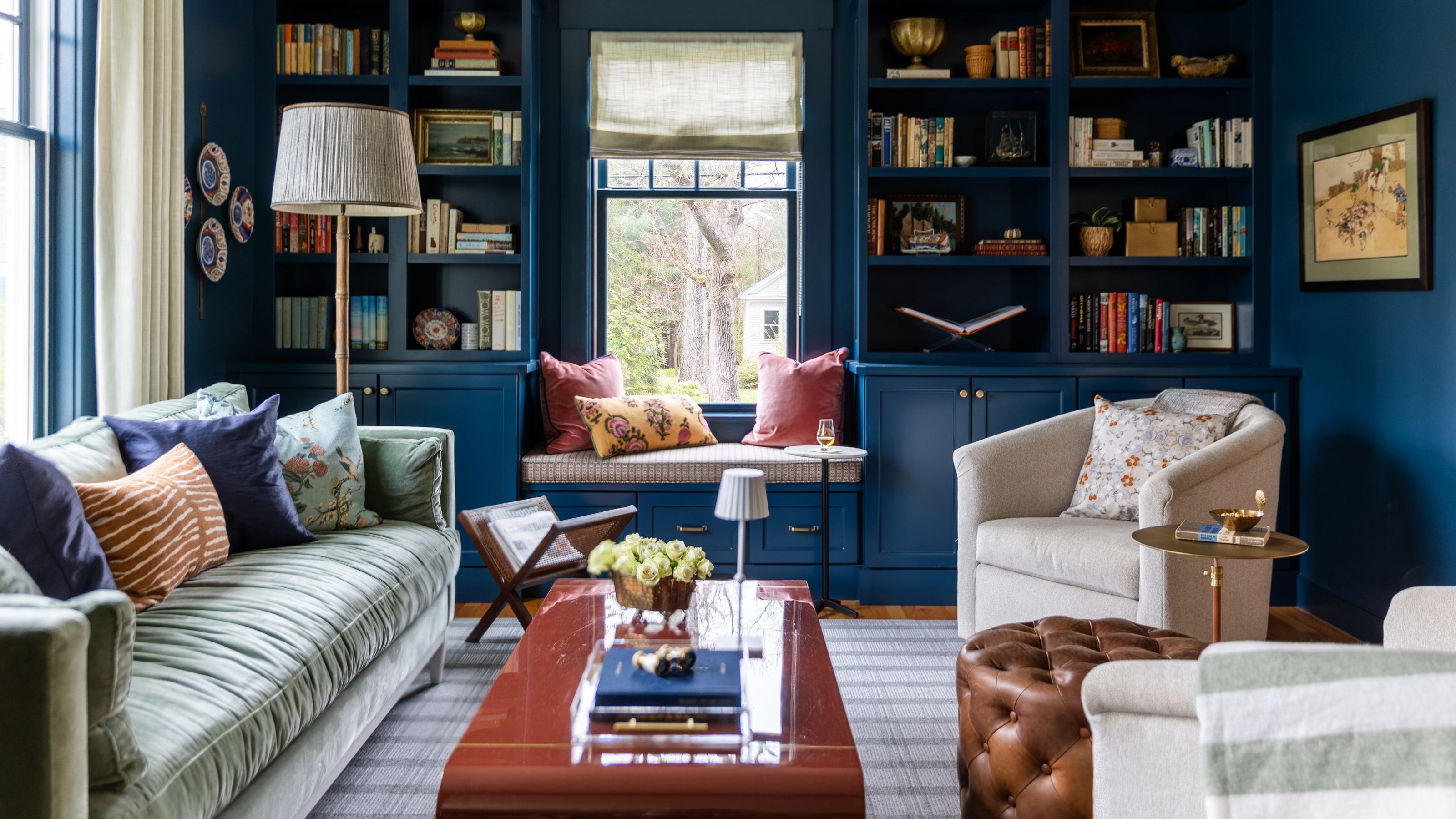 navy blue living room with custom bookcases built around a window with a nook seat, a velvet green couch, glossy coffee table and neutral tub chairs
