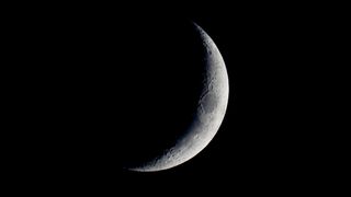 An image of the waxing crescent moon pictured against the dark sky. The lunar sea Mare Crisium can be seen as a dark oval marking the lit section of the moon's surface.