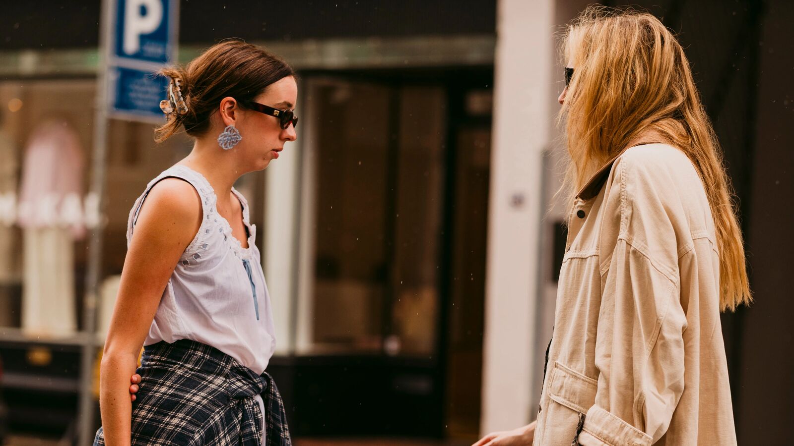 Two women are chatting wearing cute pics. One is wearing a dress over khaki trousers wit a plaid shirt around the waist. The other is wearing a barn jacket, pencil skirt, and mules. 