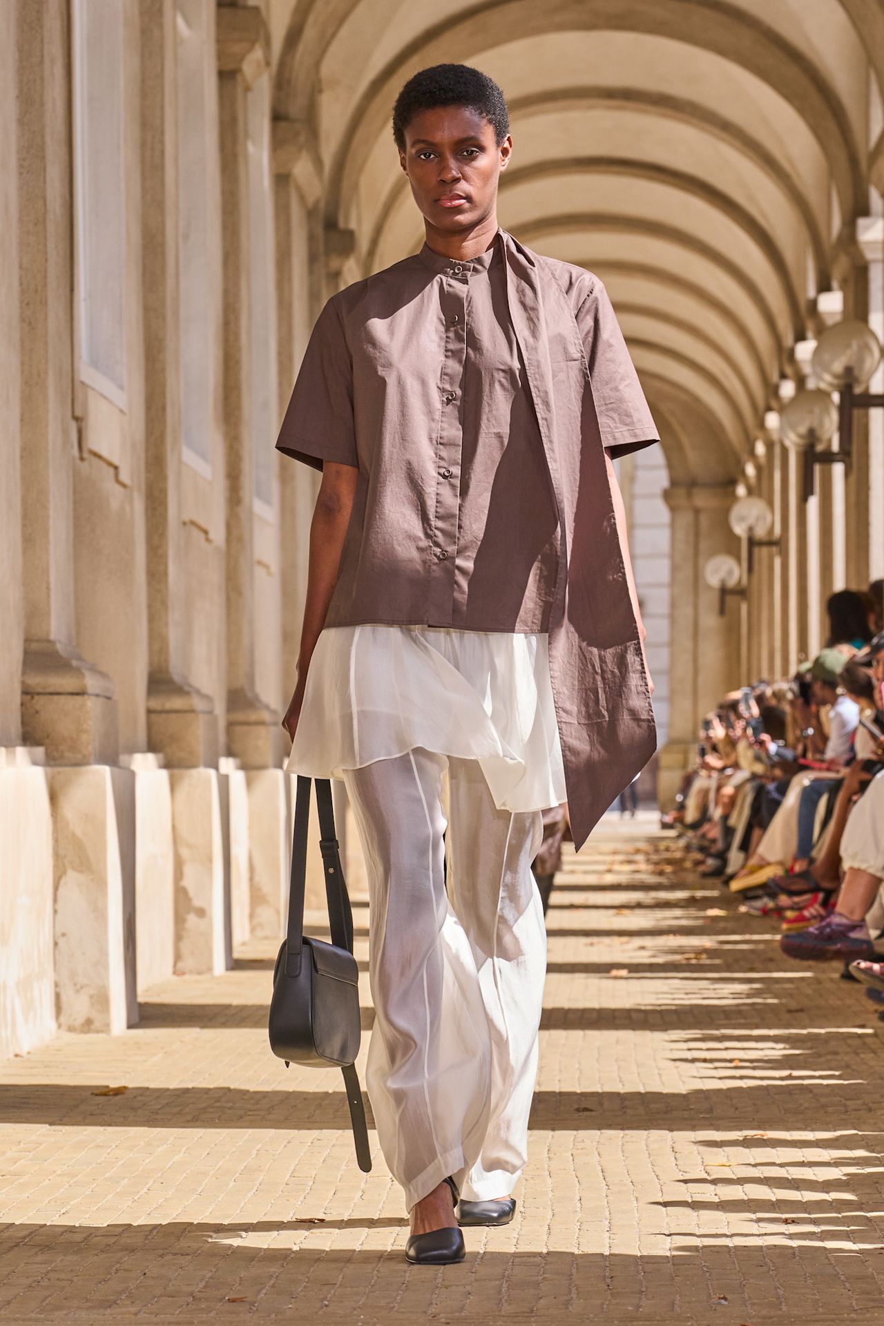 Layering staples for spring 2026 is shown in a photo from The Garment's runway show, with a model walking while wearing a cap sleeve tan button-down shirt styled on top of a sheer white slip skirt on top of matching sheer organza trousers that's paired with black square toe flats and a black shoulder bag