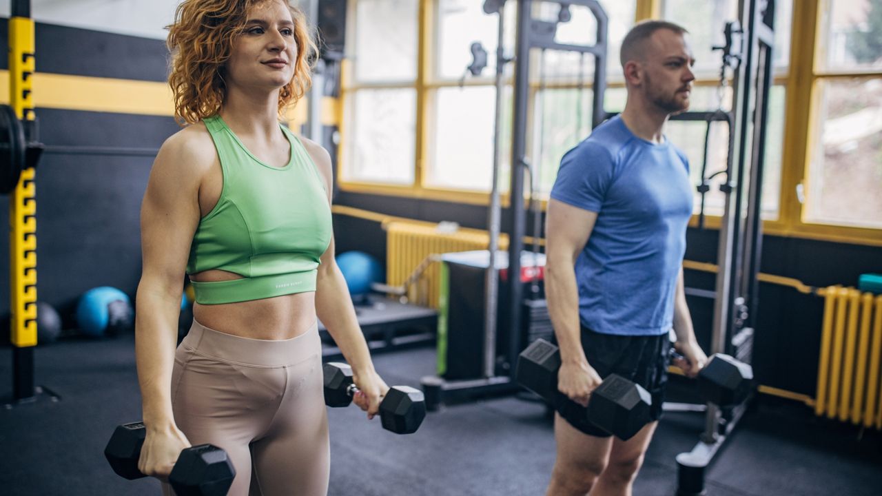 woman and man in a gym environment both standing tall holding two dumbbells one in each hand 