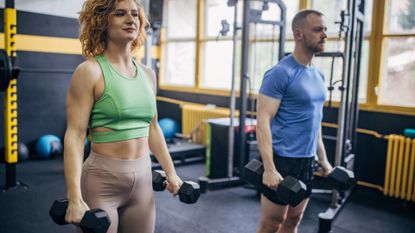woman and man in a gym environment both standing tall holding two dumbbells one in each hand