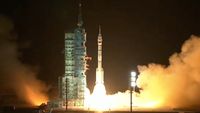 A white rocket launches next to a green tower at night, fire shooting from its bottom, a smoke plume blowing to the right.