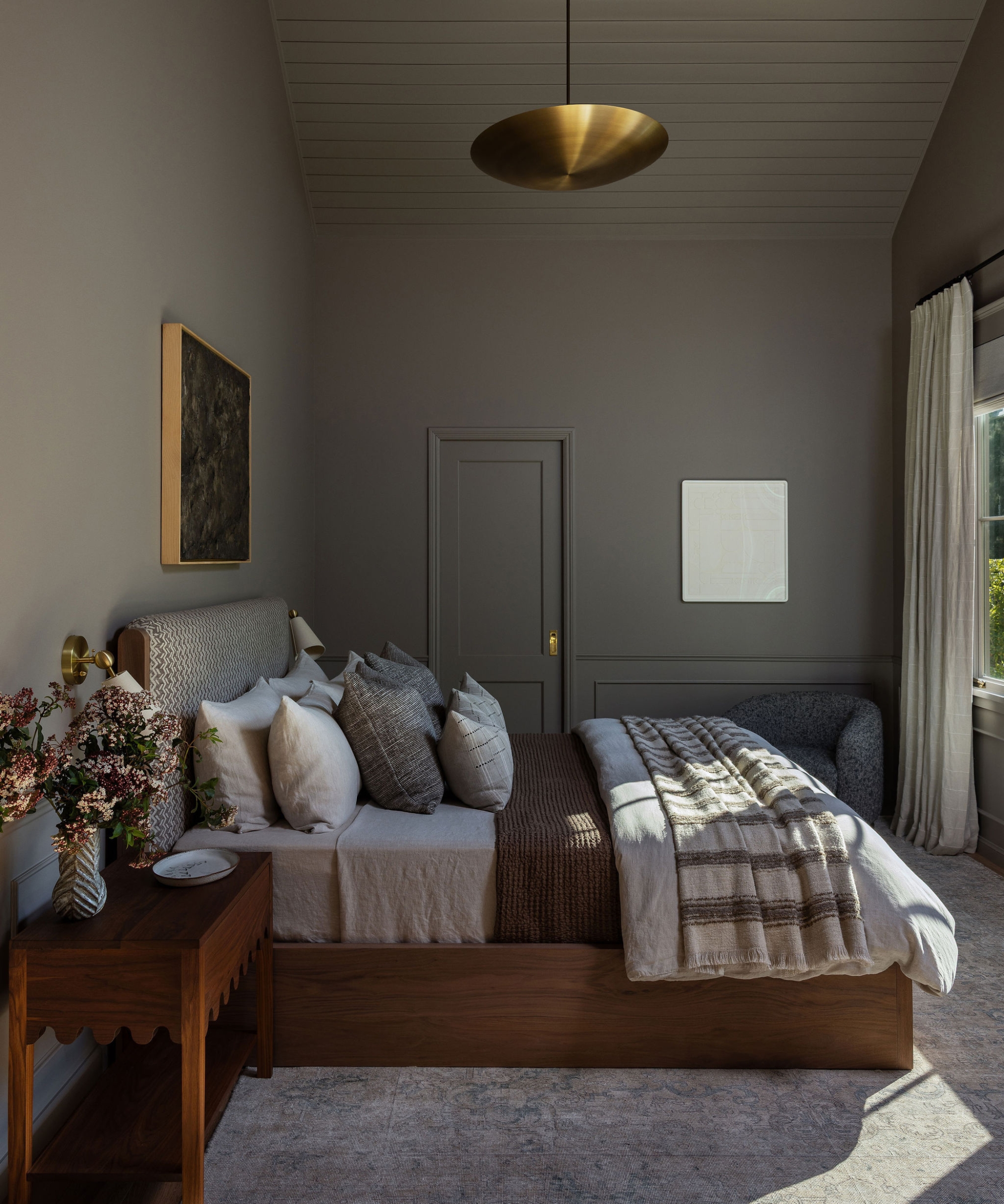Primary bedroom with gray walls, a wood bed, patterned upholstered headboard, scalloped nightstand and brass pendant light.