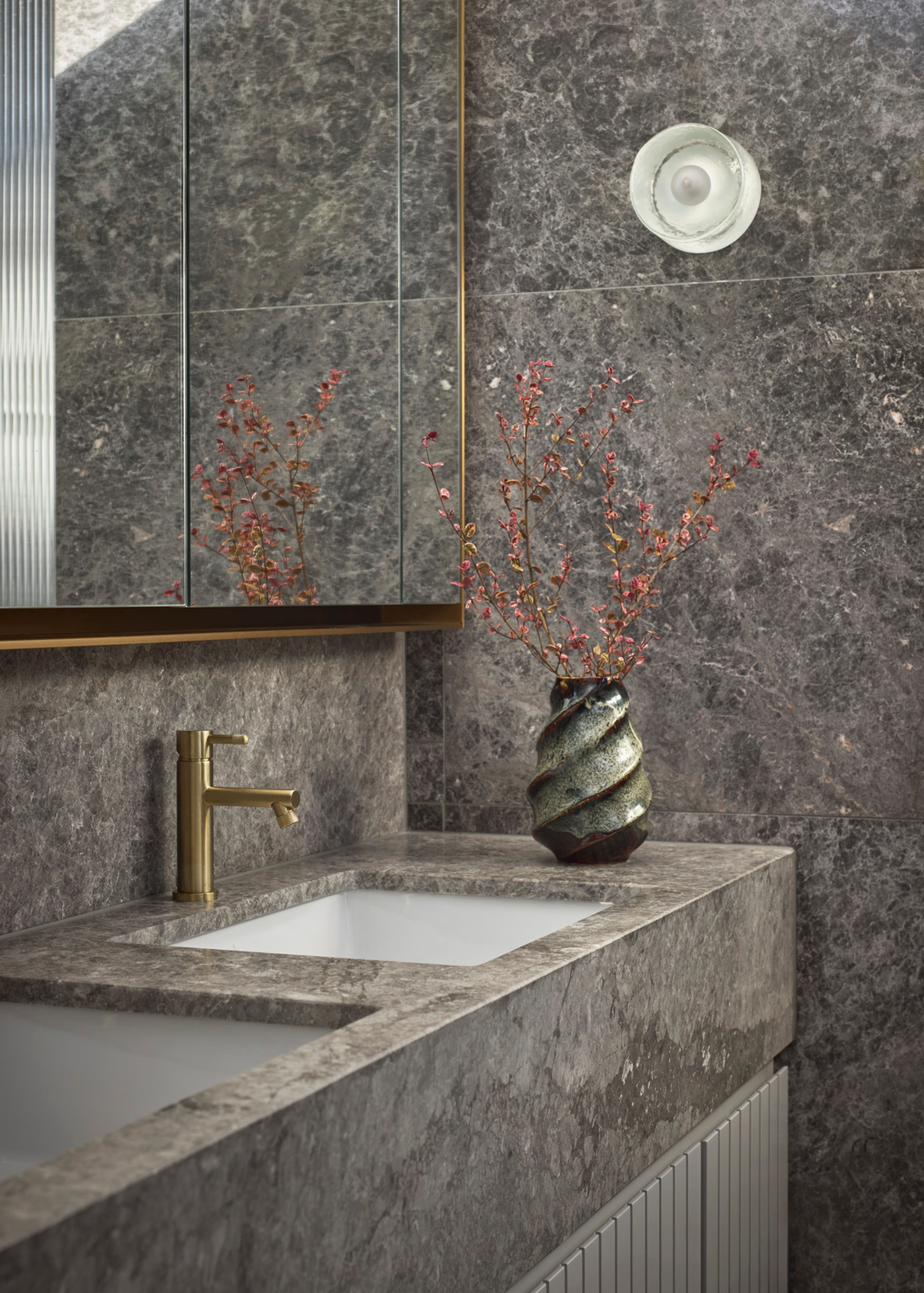 A bathroom with gray marble walls and sink-top by a swirl vase of stems and a glass wall sconce
