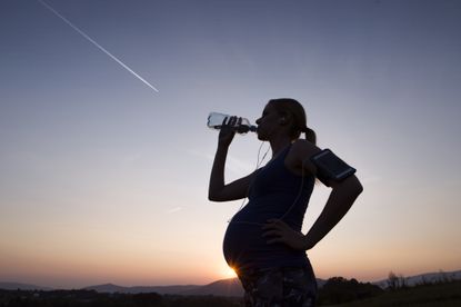 Silhouette of woman running while pregnant and drinking water from bottle at sunset