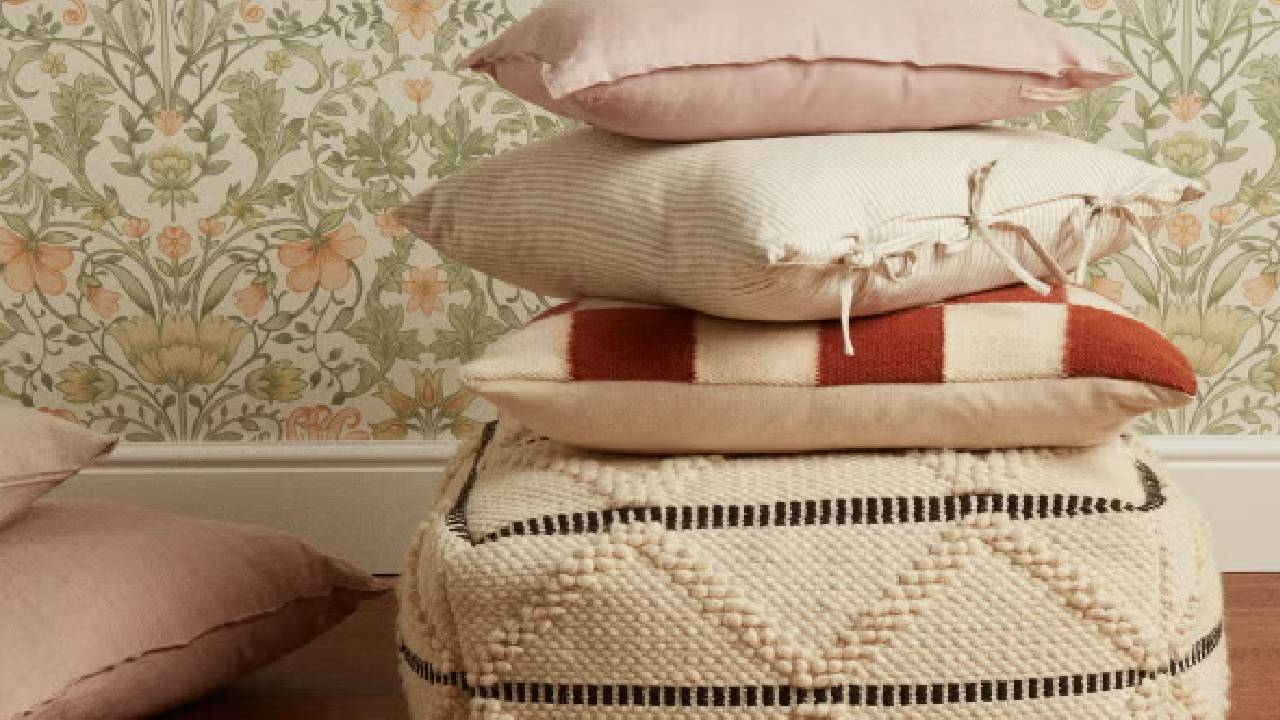 A pile of pink, white and stripy red cushions from The Hut, in front of green and pink floral wallpaper. 