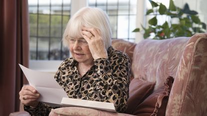 An older woman looks concerned as she looks at paperwork while sitting on her couch.