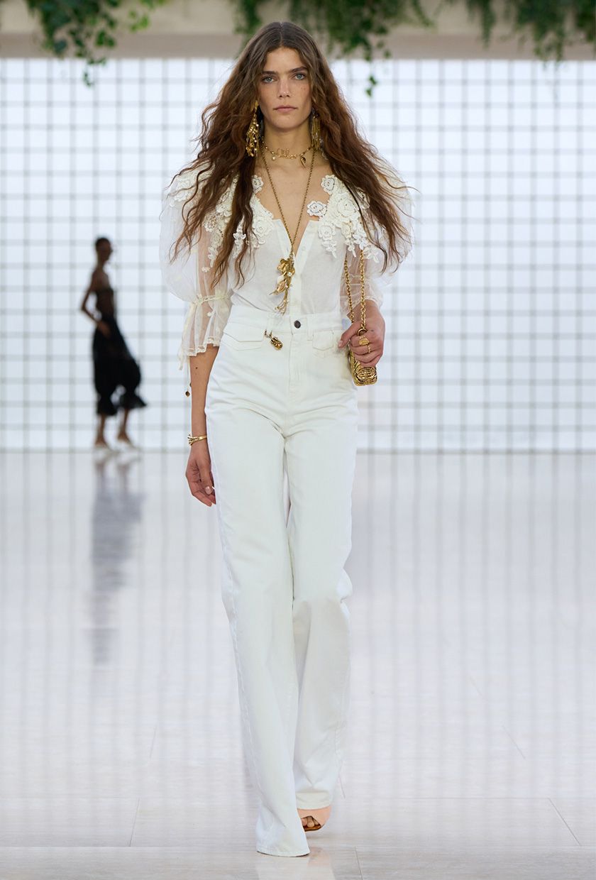 The coastal boho trend is shown in Chloé's spring summer 2025 runway show, with a model walking down the runway wearing long gold earrings and a gold lariat necklace styled with a white lace puff-sleeve top, high-waisted wide leg jeans, nude sandals, and a gold bag