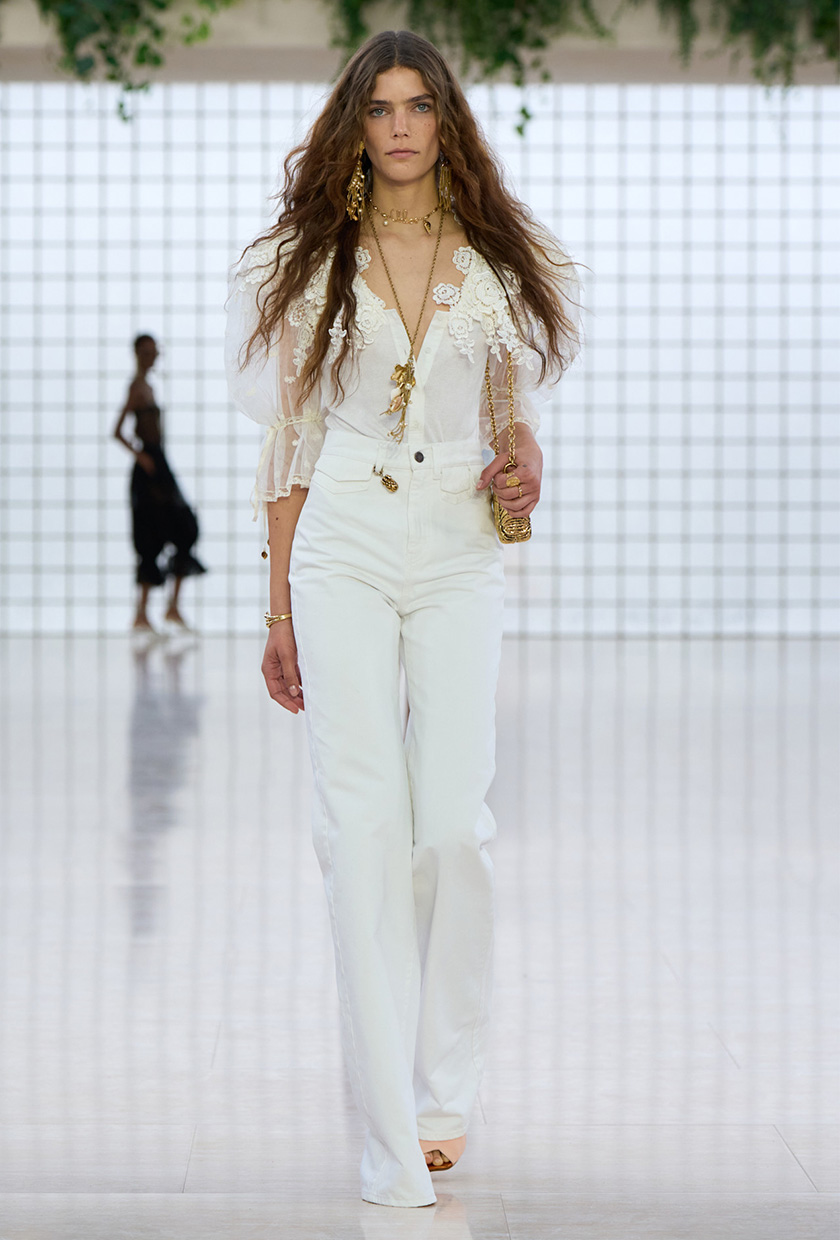 The coastal boho trend is shown in Chloé's spring summer 2025 runway show, with a model walking down the runway wearing long gold earrings and a gold lariat necklace styled with a white lace puff-sleeve top, high-waisted wide leg jeans, nude sandals, and a gold bag