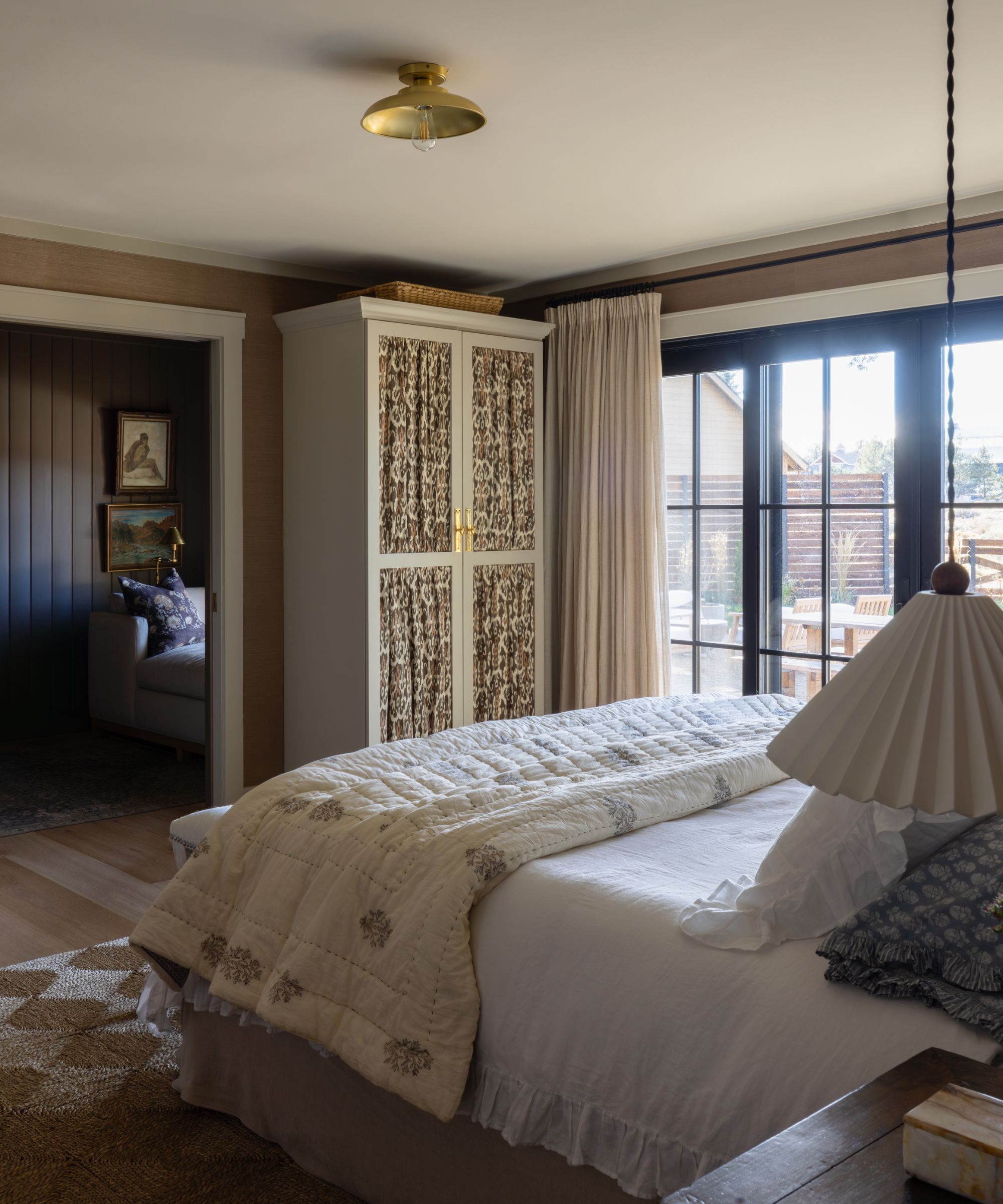 Calm bedroom with white bed sheets, pleated lamp, small wardrobe with floral fabric on the door, and large windows