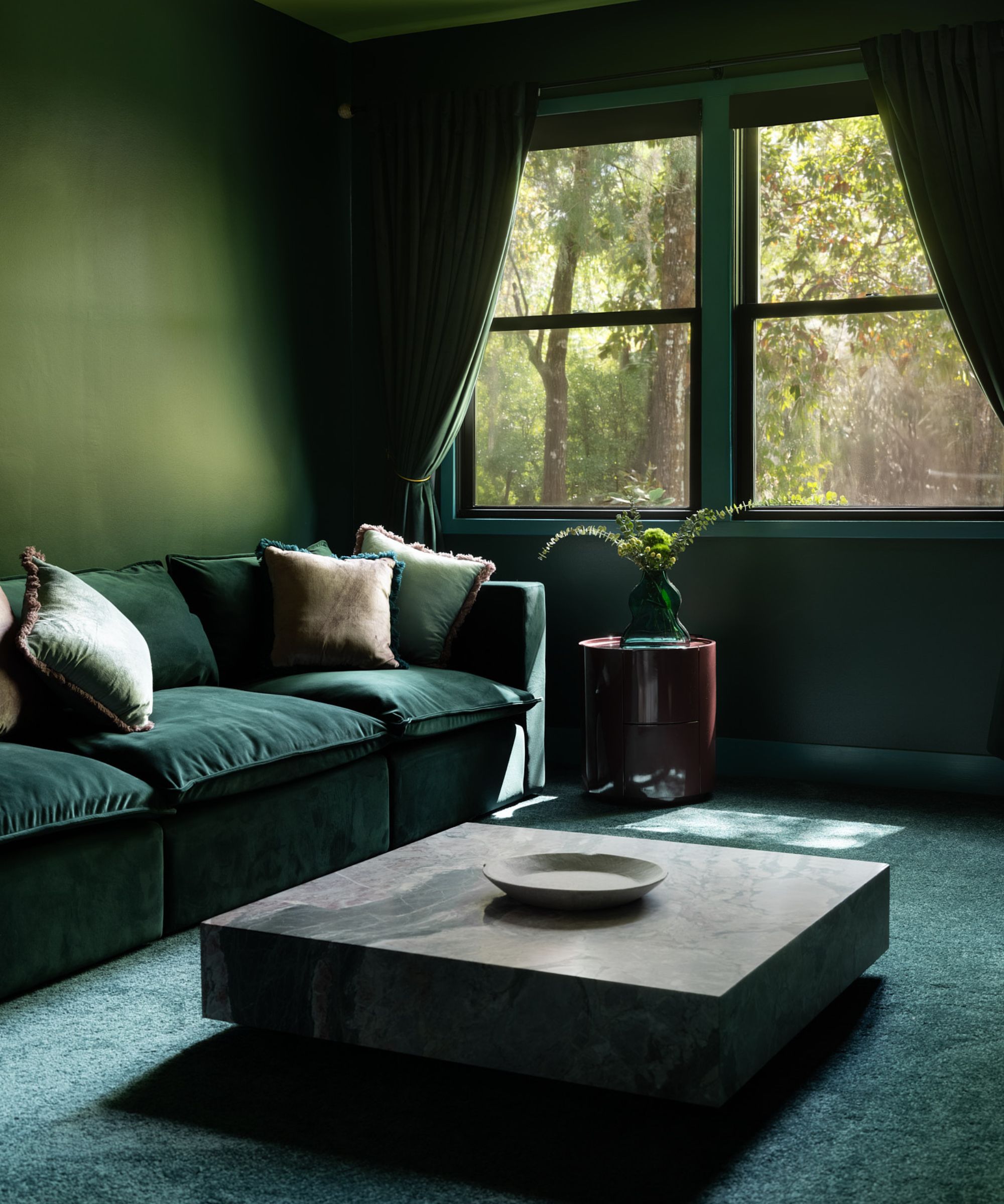 A cozy and modern snug room with dark green walls, a teal carpet, a forest green sofa, a low coffee table, and windows with dark green curtains.