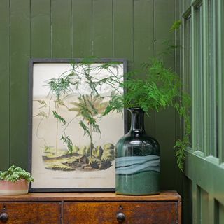 An old wooden chest of drawers with a green vase and foliage on top and green painted wood panelling on the walls