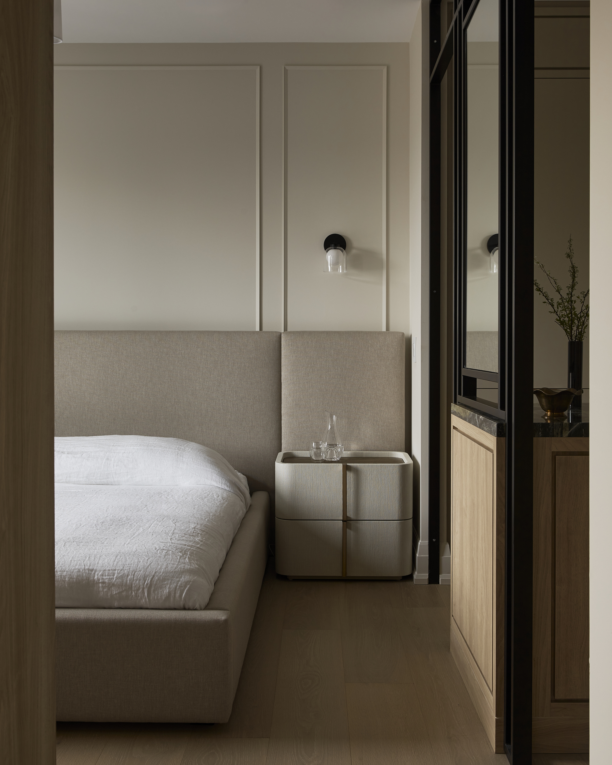 modern bedroom with neutral palette, upholstered bed frame with extended headboard, sconce light on wall, beside table, and glass partition leading to closet