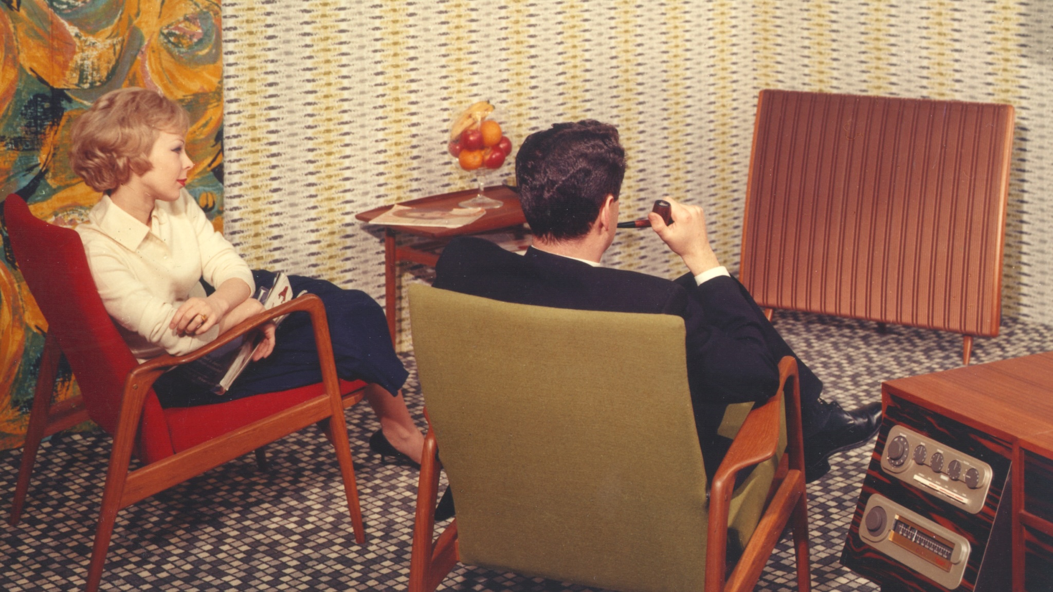 Quad ESL promotional photo from the 1950s with a seated couple on separate chairs looking at the speaker in their front room. The man is smoking a pipe.