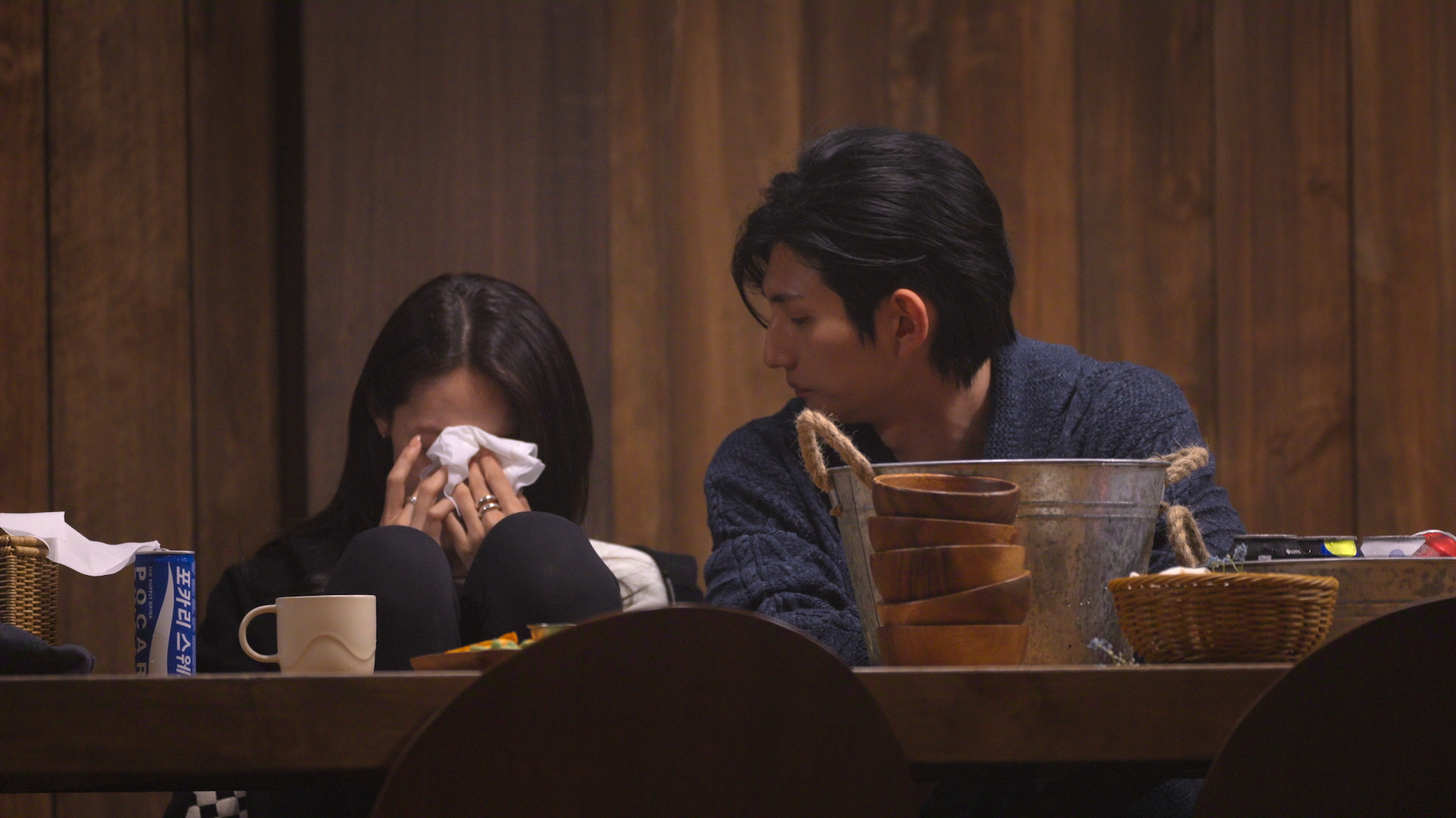 A woman (Mina Sue Choi) covers her face with a tissue while sitting next to a man (Samuel Lee) wearing a navy sweater. They sit behind a wooden table holding a tissue box, mug, silver bucket, and small straw baskets, in a still from 'Single's Inferno' season 5.
