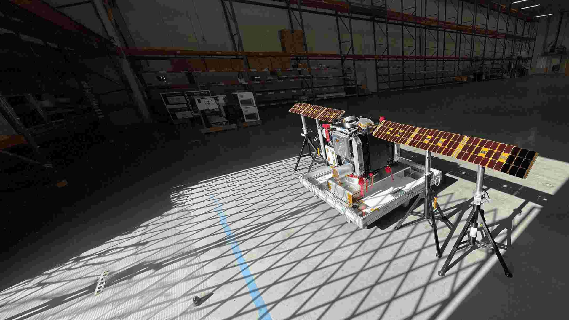 A metal machine with two outstretched solar panels sits in an empty warehouse