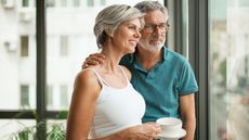 An older couple stand together at a window looking out.