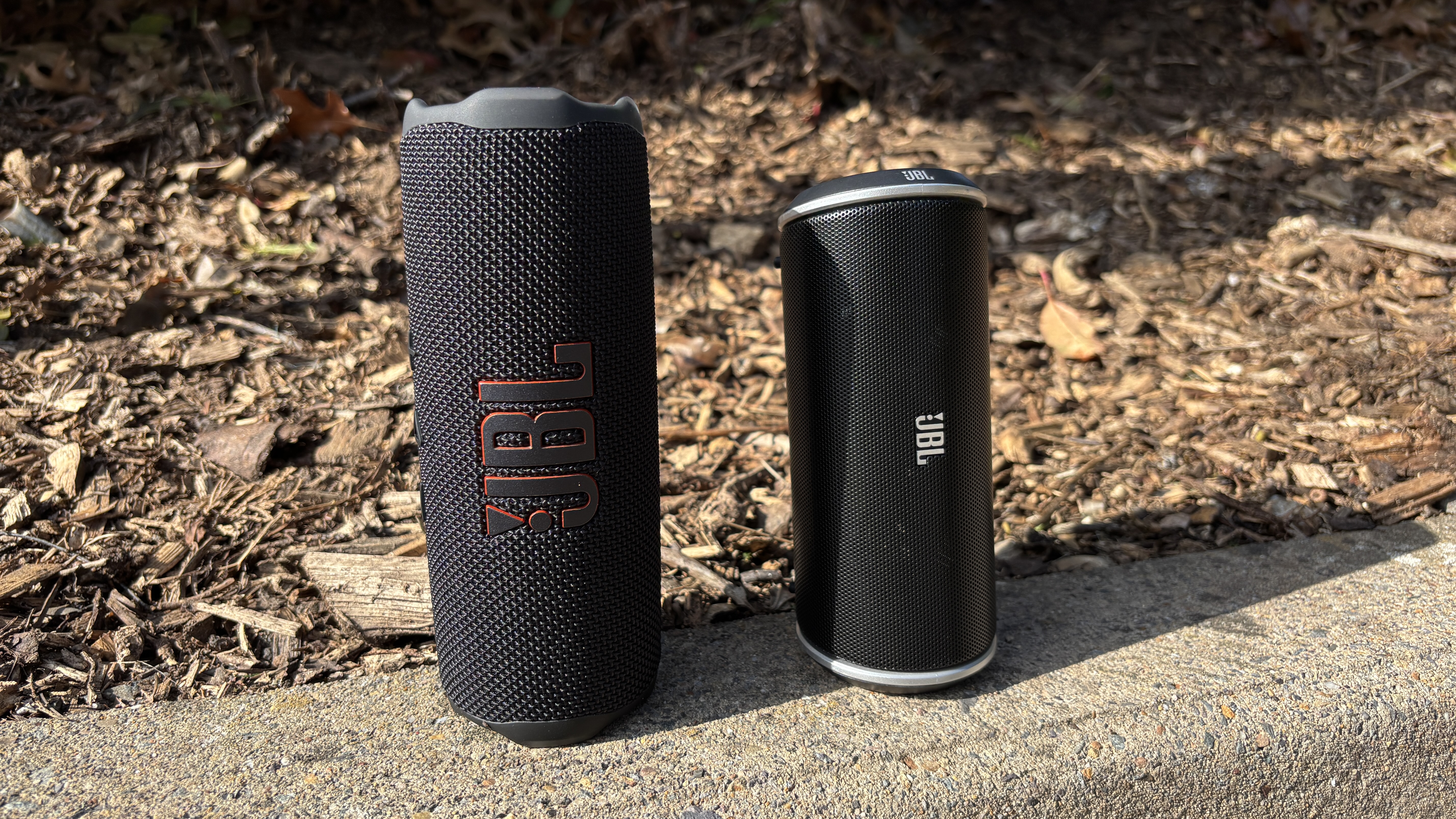JBL Flip 7 stood next to the original JBL Flip on the edge of a kerb