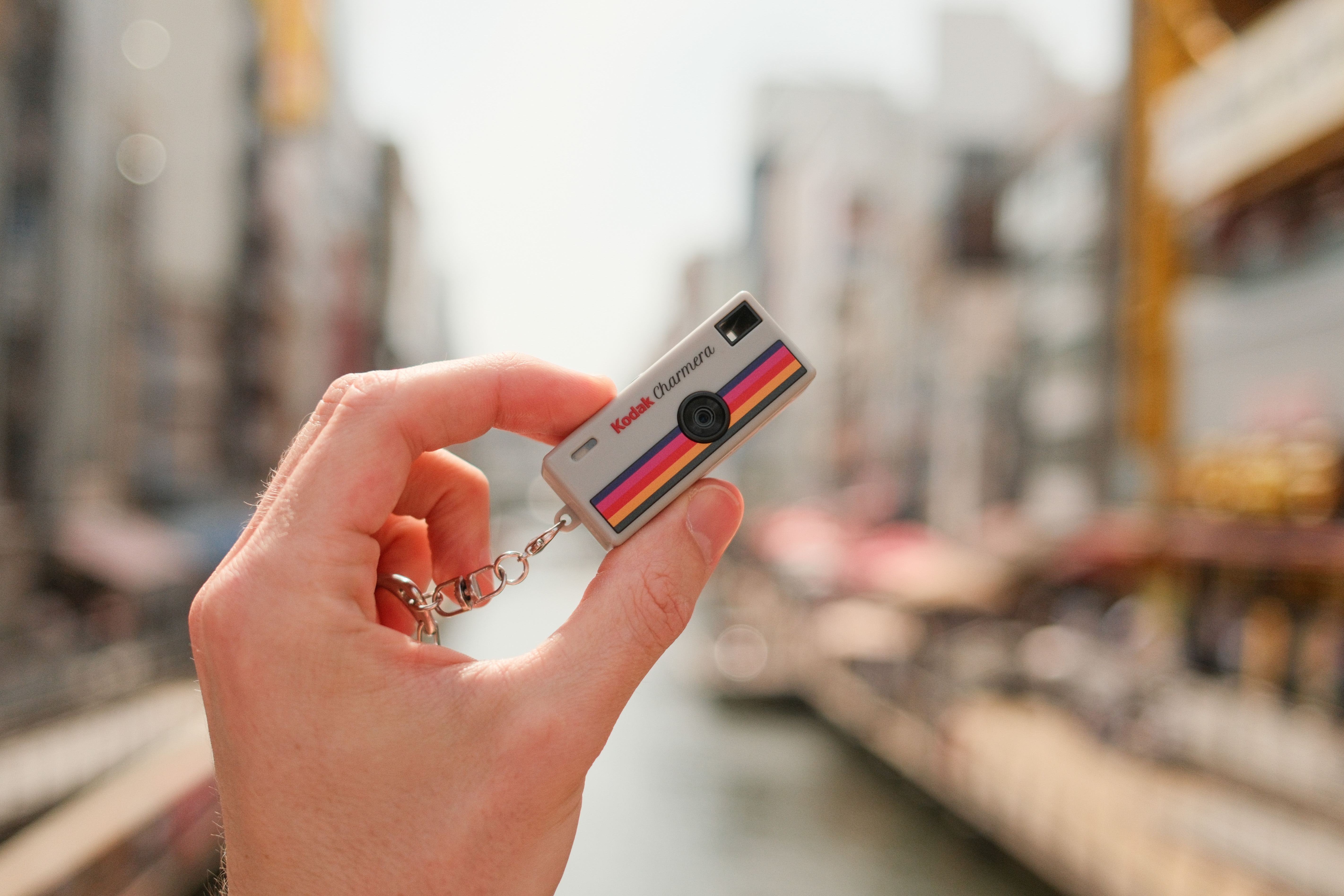 Hand holding a tiny Kodak Charmera keychain camera against a blurred canal background.