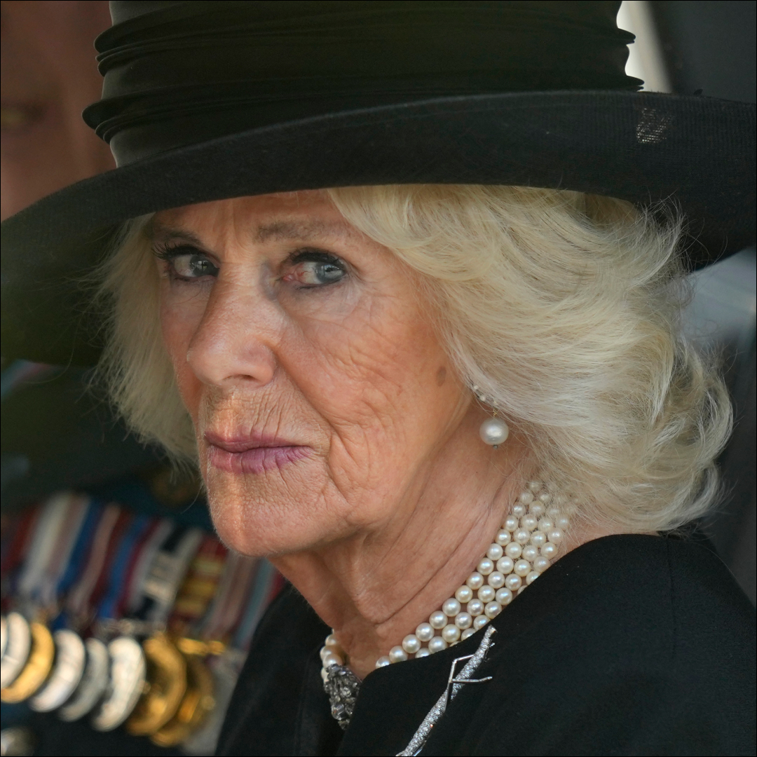 Queen Camilla wears a black hat and black dress with a string of pearls