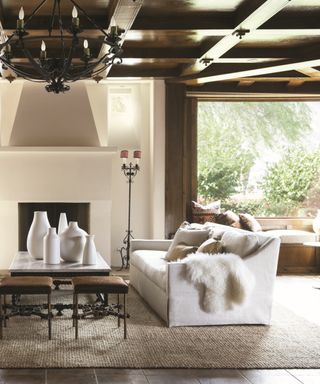 neutral rustic living room with beams, tiled floor, large jute rug, light gray couch, sheepskins, white vases, coffee table, stools, iron chandelier, fireplace, creamy walls, window seat