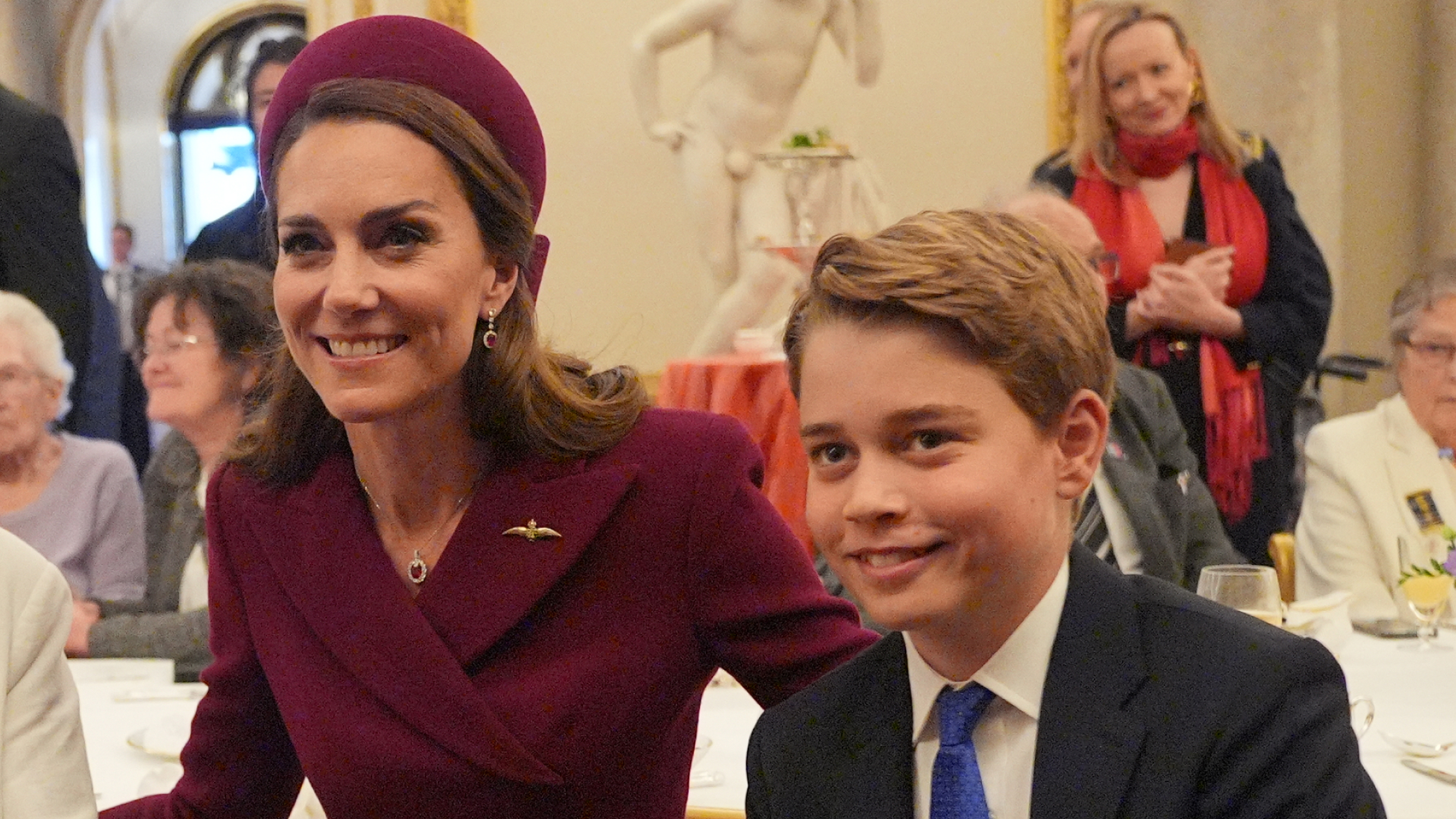 Kate Middleton wears a burgundy coat and matching hat and poses with son Prince George, who is wearing a navy suit, white shirt, and blue tie