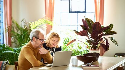 A couple sits at their kitchen table looking at a laptop.