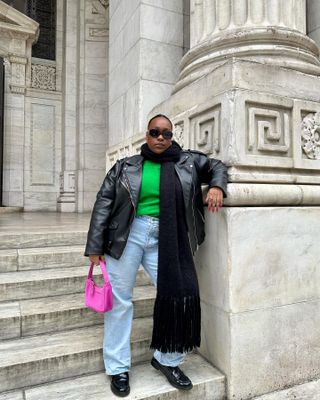 Woman in leather jacket, green sweater, light-wash jeans, and loafers with a pink bag