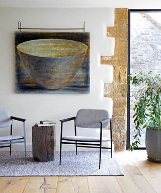 Two matching armchairs either side a stump of wood acting as a side table, artwork on wall, exposed brickwork, plant, large window