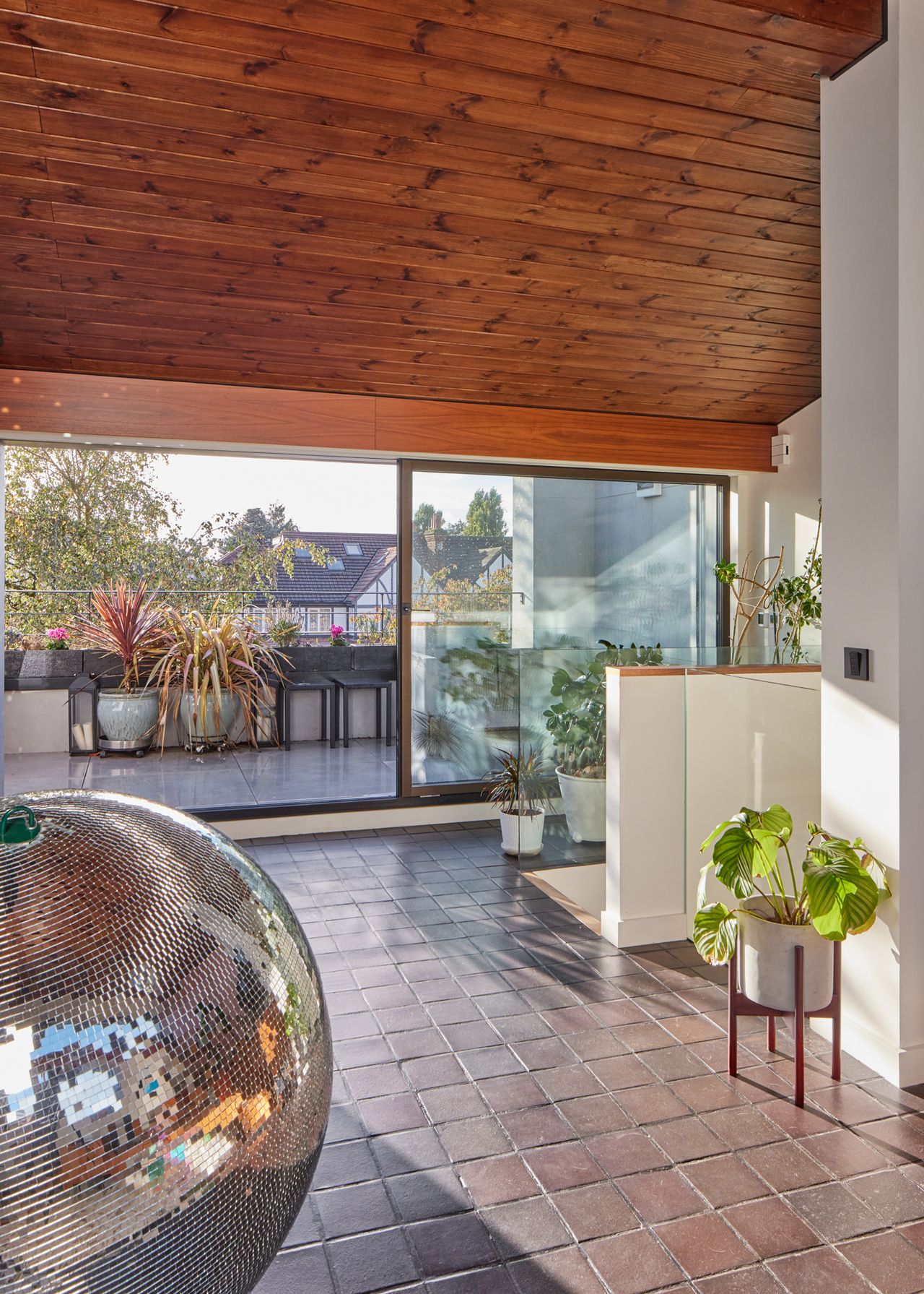 The room of a modernist home with a wood panelled ceilings, tiled floors, houseplants and a large disco ball. Door are open, leading out to an outside terrace