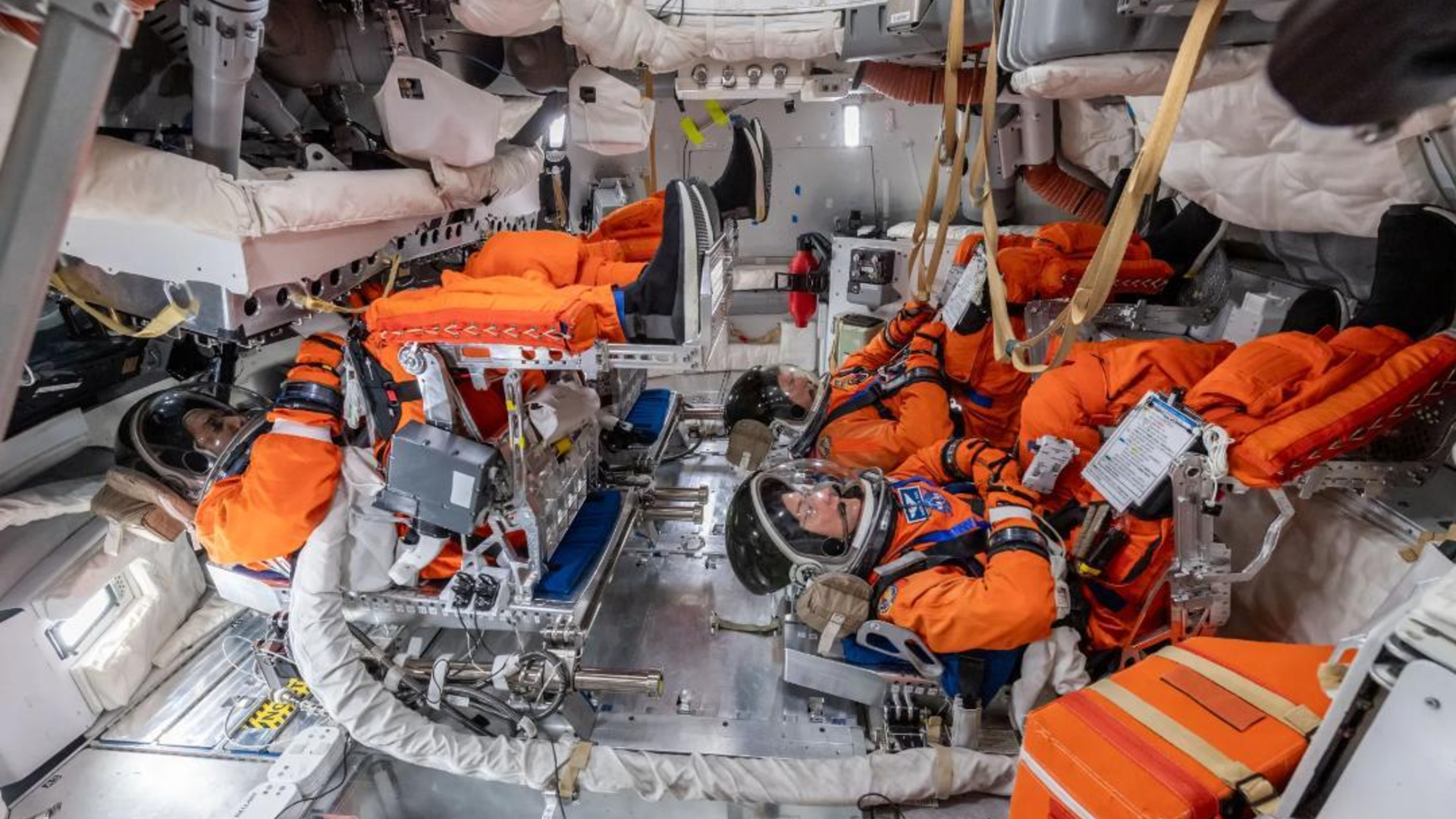Four individuals wearing orange space suits and clear helmets are buckled into the floor of a full spacecraft
