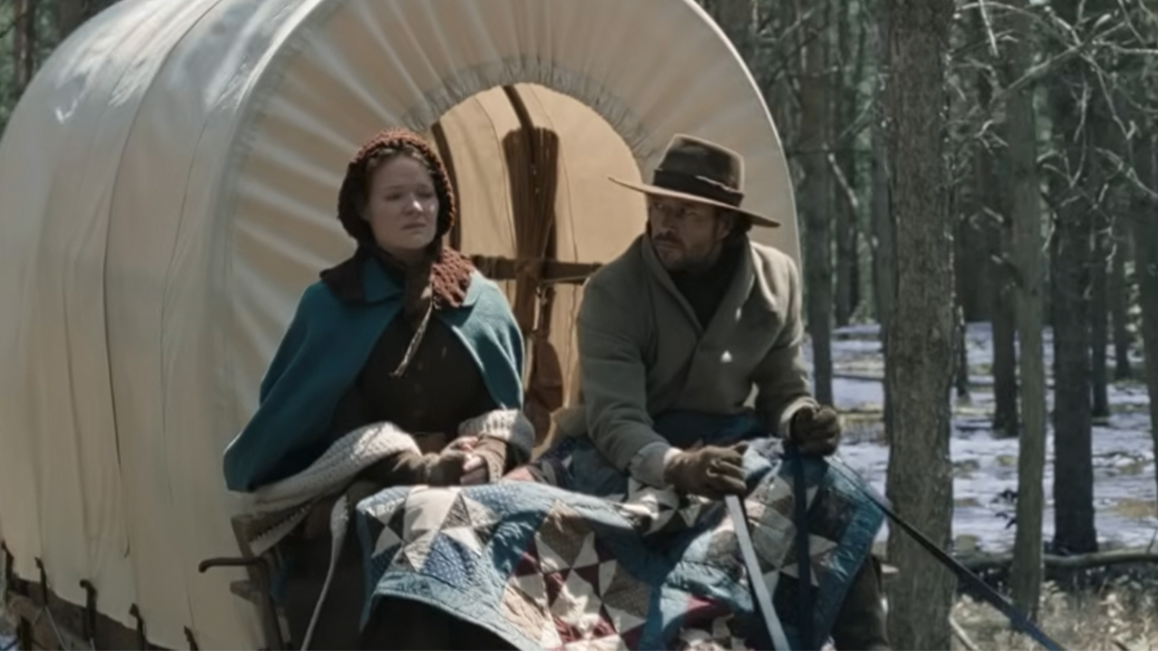 Two people traveling in the front of a covered wagon in Little House on the Prairie. 