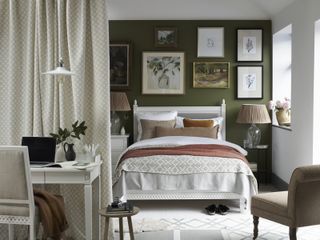 A bedroom with a dark green wall, a white bed and a dressing table that doubles as a desk