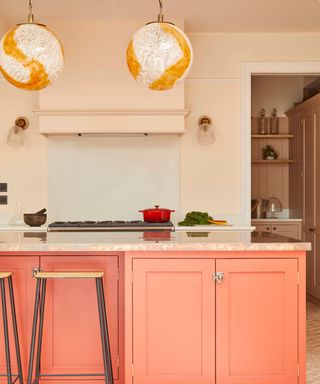 A pink kitchen with two white and orange globe pendant lights, light pink walls, and a coral colored kitchen island with two stools next to it