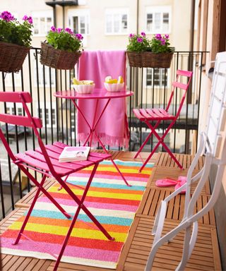 Small colorful balcony with striped rainbow rug pink metal bistro set and hanging planters with purple pansies