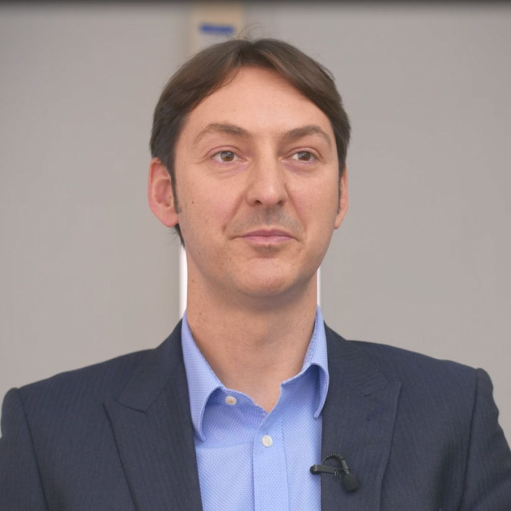 A professional headshot of a man with short dark hair wearing a navy blazer and light blue shirt, with a lapel microphone, against a grey background.
