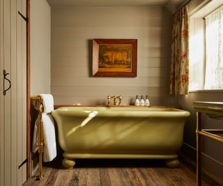 Olive green freestanding bath with brass taps and towel rail against panelled wall, framed cattle print.