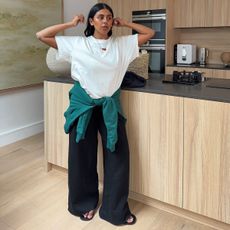 @monikh wears a pair of black wide-leg trousers with a white T-shirt and a teal jumper around her waist