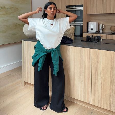 @monikh wears a pair of black wide-leg trousers with a white T-shirt and a teal jumper around her waist