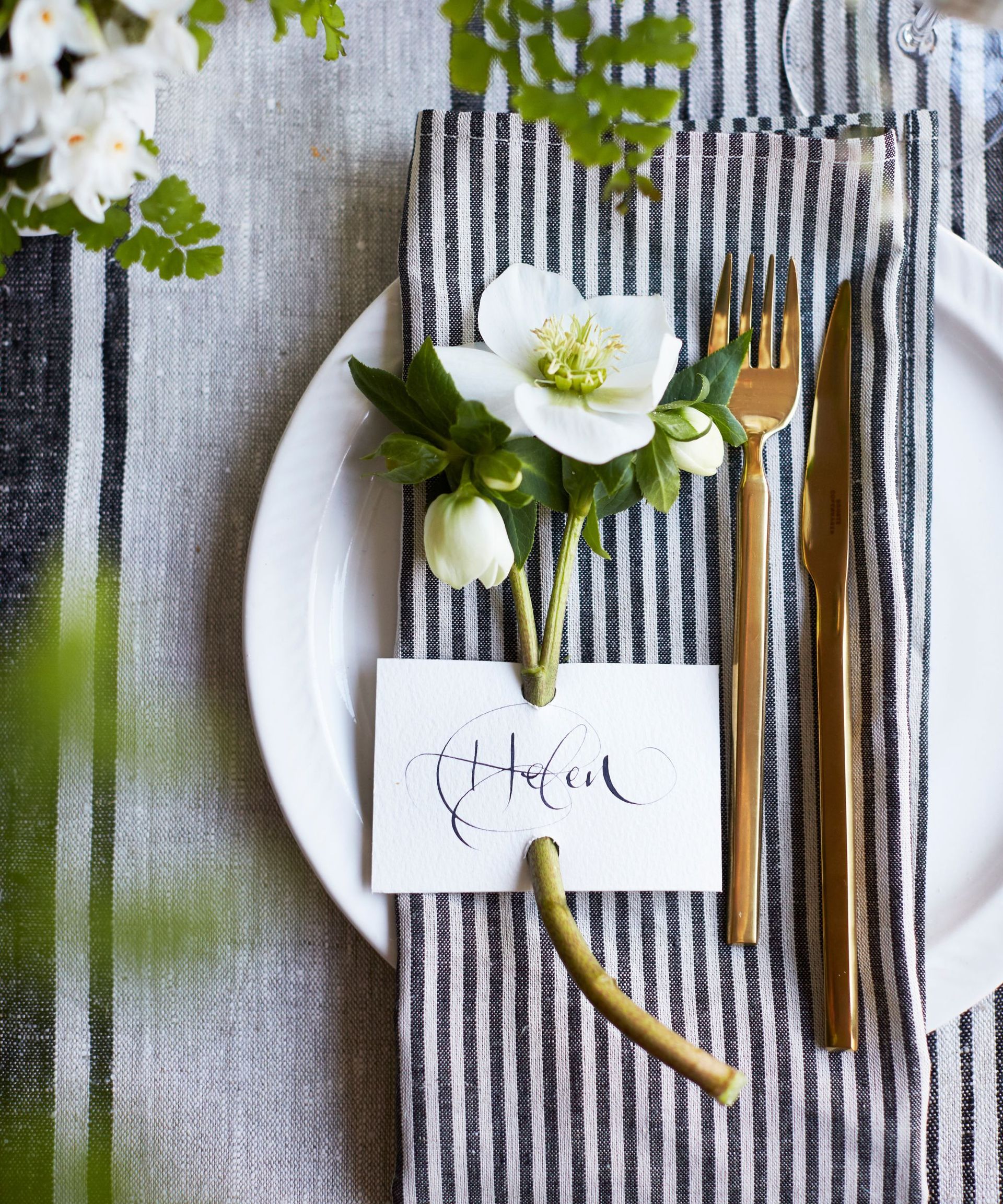 White place card with calligraphy writing attached to a white flower on a navy and white striped napkin next to a set of gold cutlery