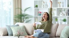 Excited woman celebrating with her fist in the air while holding a bill and sitting on the couch at home